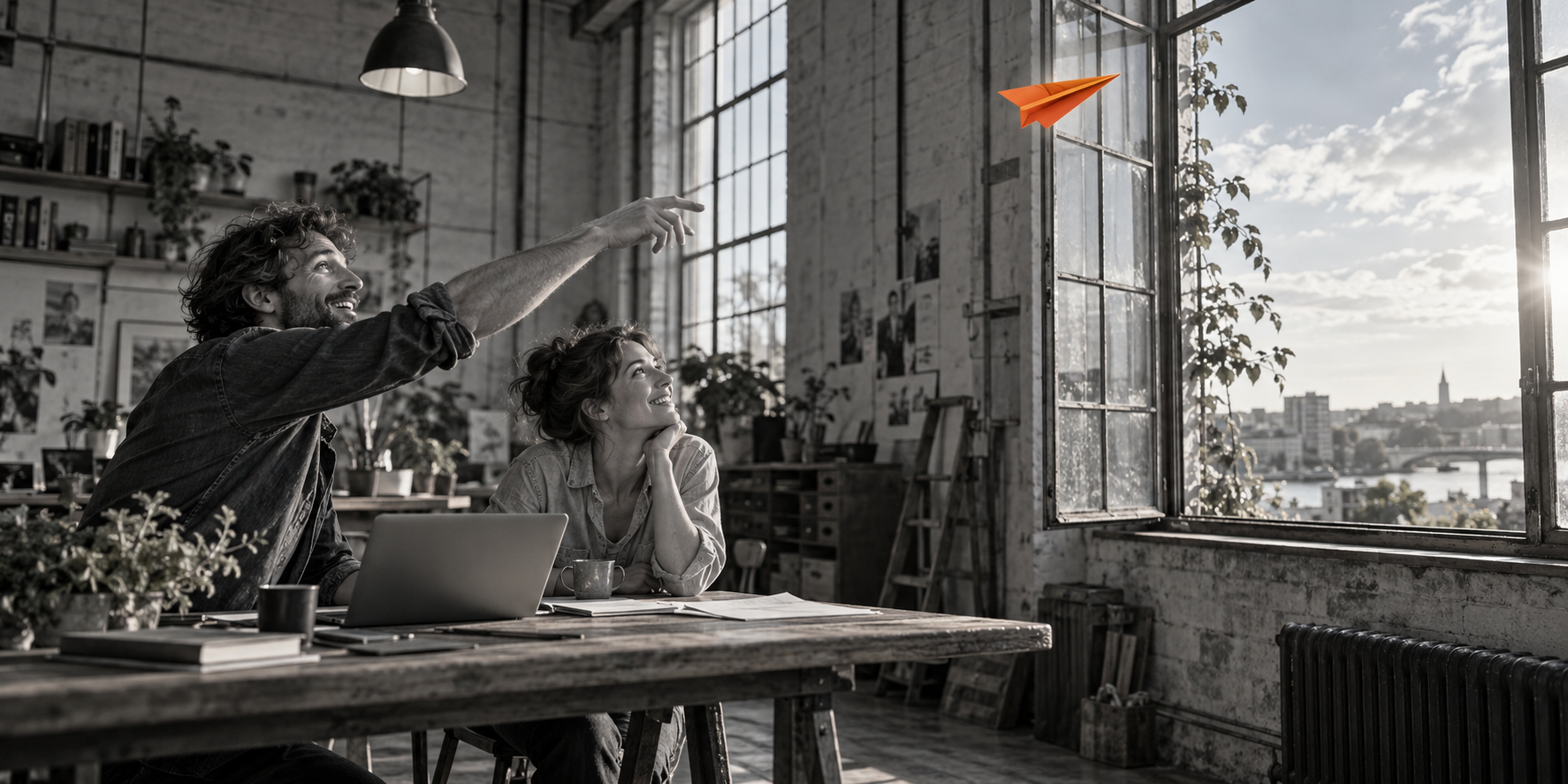 Two people in a monochrome workshop watch an orange paper glider fly towards an open window.
