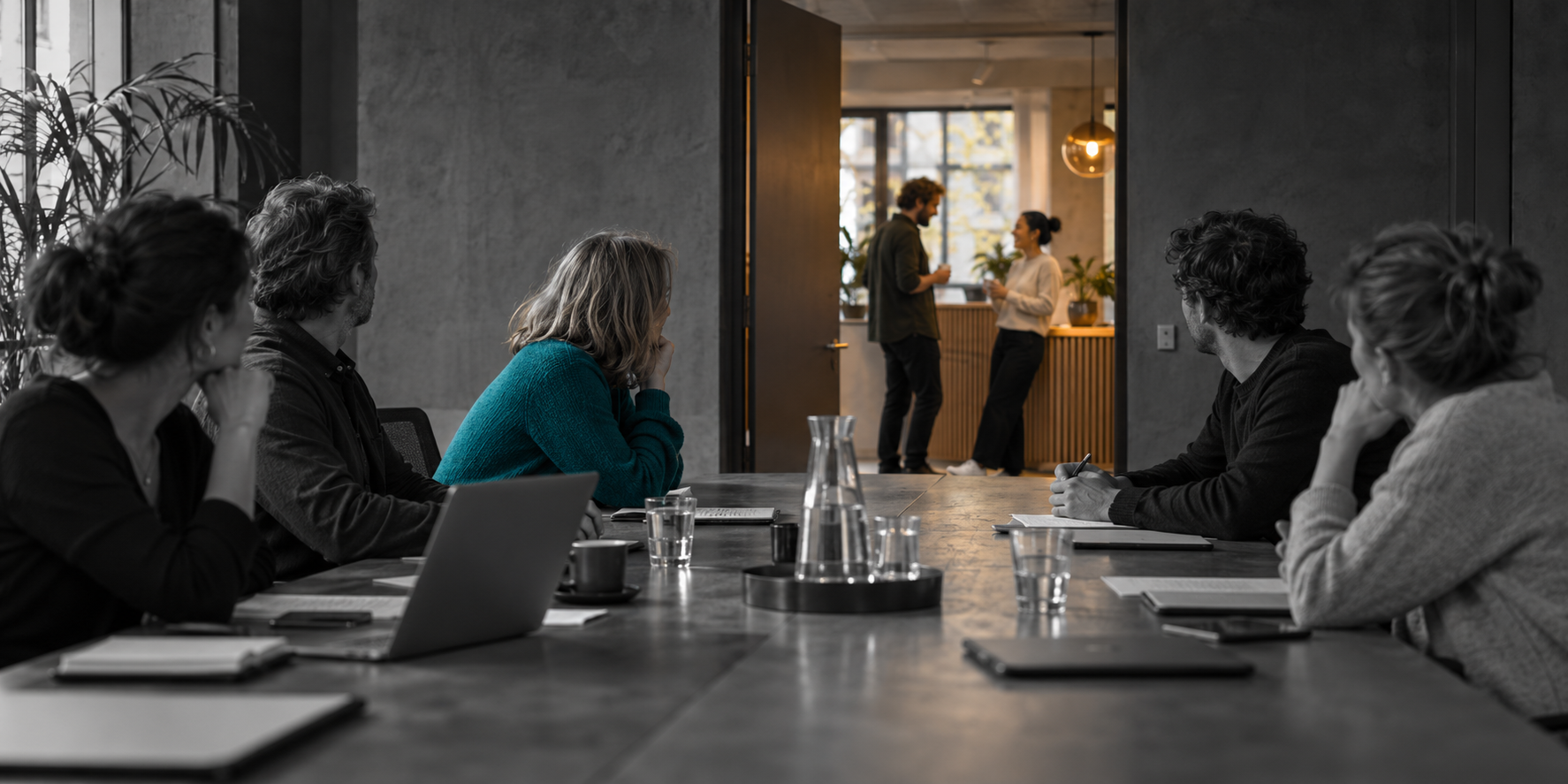 People in a monochrome boardroom turn towards a conversation outside an open door.